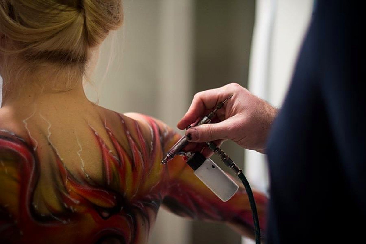 Dwight Pepper airbrushing a flowing red and gold design onto a model's back, airbrush in hand
